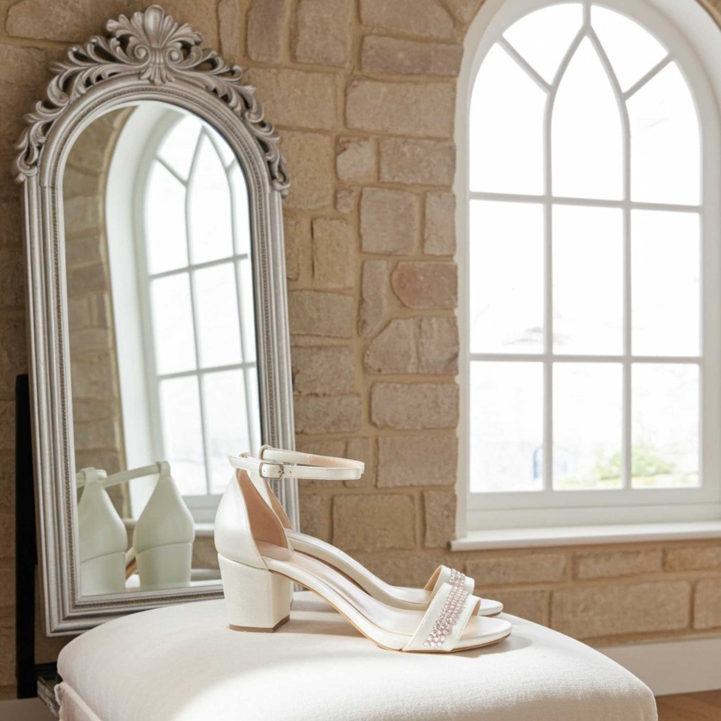 Ivory block heel shoes on a white stool in front of a decorative mirror with a stone wall background.