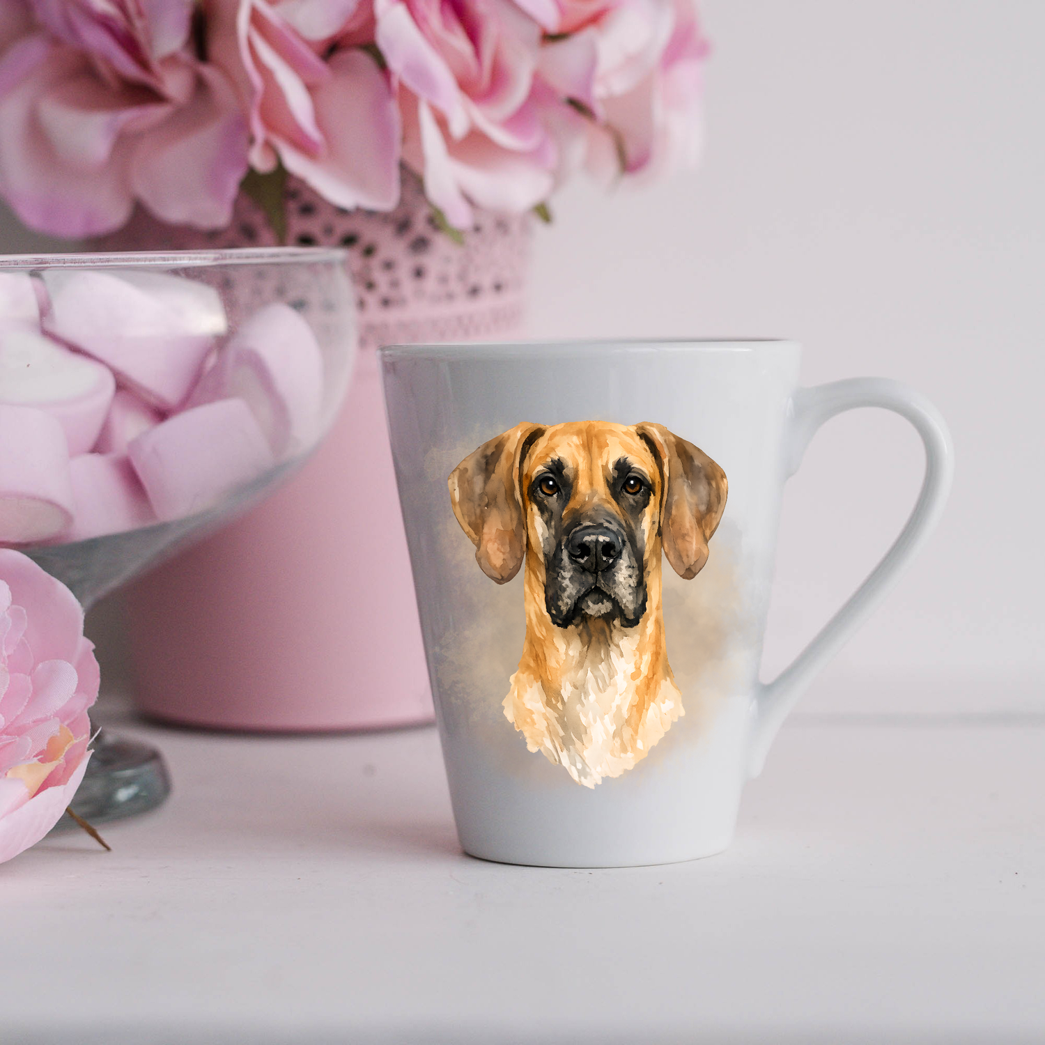A Fawn Great Dane head-shot is placed on a white ceramic latte mug in a smoky background and looking forward.  The mug is placed on a white surface with pink flowers in a pink plant pot and a dish of pink marshmallows.