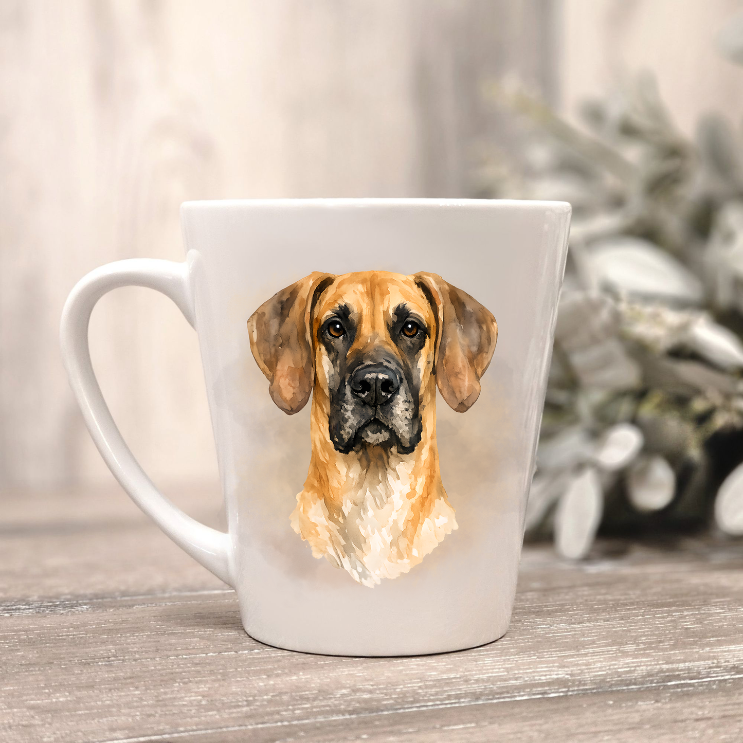 A Fawn Great Dane head on a white ceramic latte mug in a smoky background and looking forward.  The mug is placed on a wooden surface with sage green foliage to the right-hand side.