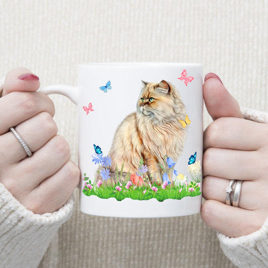 A woman is holding white ceramic mug showing a light coloured Persian cat sitting on grass with flowers and watching butterflies.