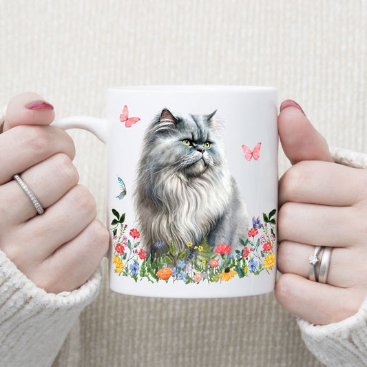 A white ceramic mug with a Persian Cat with long grey hair and green eyes is sat among multi-coloured wildflowers and watching pink butterflies. A woman wearing rings is holding the mug in two hands.
