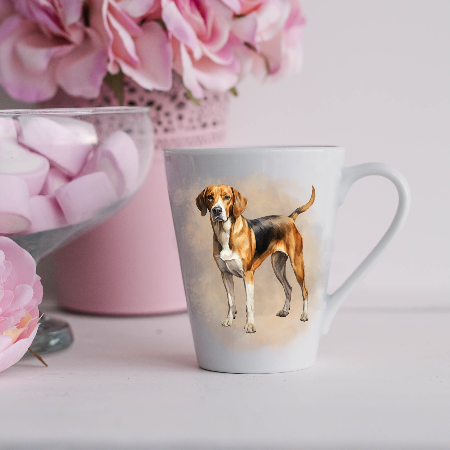 A Foxhound is shown in a standing position on a white ceramic latte mug in a smoky background and looking forward.  The mug is placed on a white surface with pink flowers in a pink plant pot and a dish of pink marshmallows.