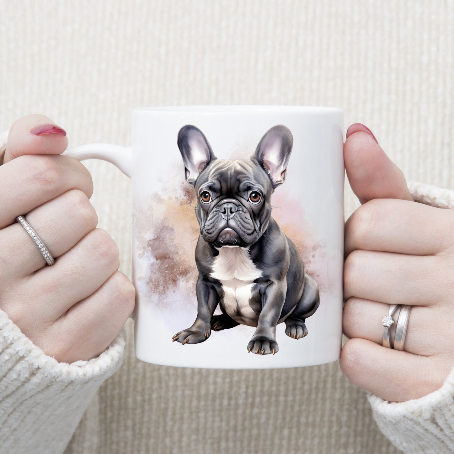 A watercolour image of a grey French Bulldog with a white chest sitting in front of a brown and gold misty background.  A woman is holding the mug in two hands.