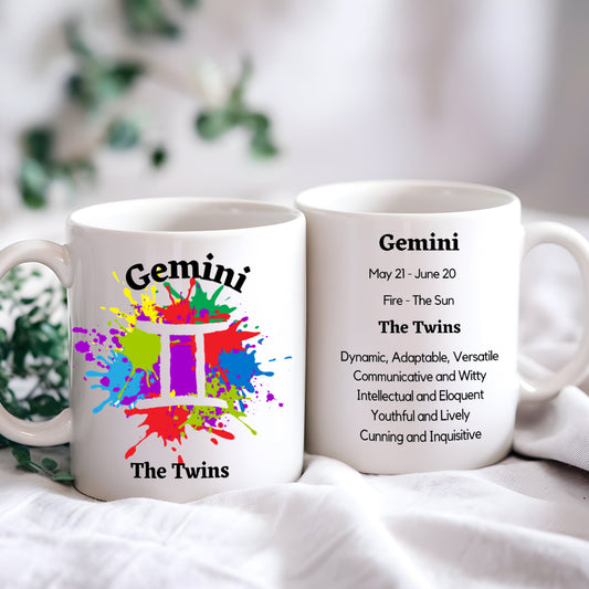 Two white ceramic coffee mugs with colorful astrological Gemini symbol and related information printed on them, placed on a surface with a plant in the background.