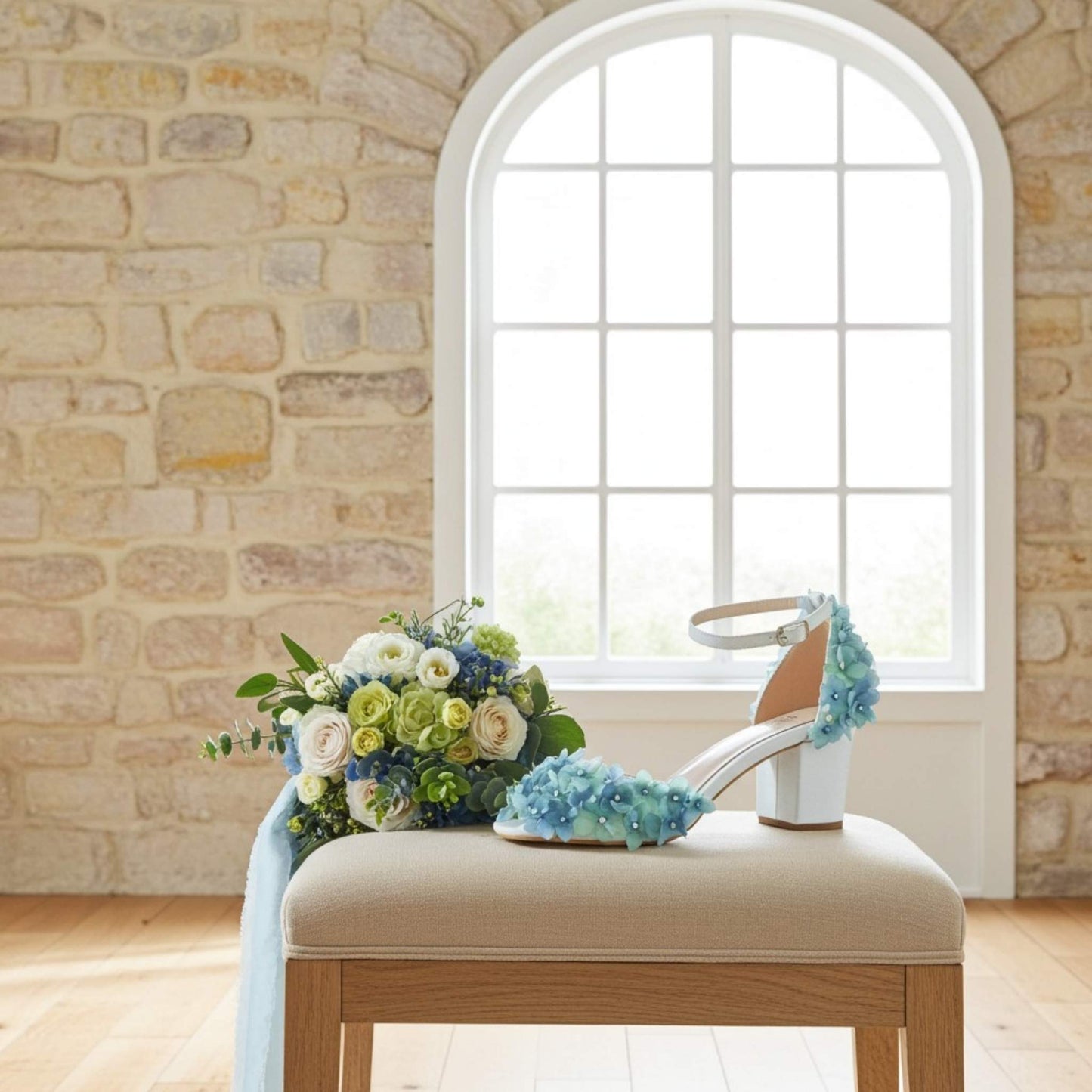 Bouquet of flowers and an ivory bridal sandal with blue floral design on a beige cushion in front of a stone wall and window.