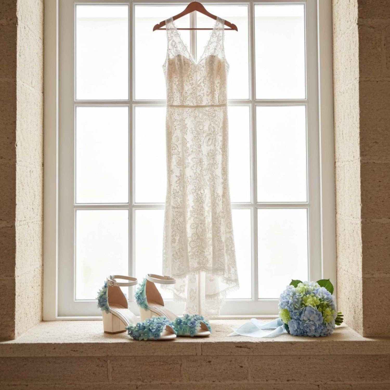 Wedding dress hanging by a window with shoes and bouquet on a windowsill.