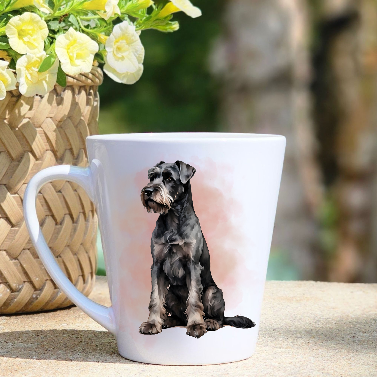 A white ceramic latte mug featuring a Giant Schnauzer in a pinky mist background.  A basket containing yellow flowers sits to the left-hand side. 