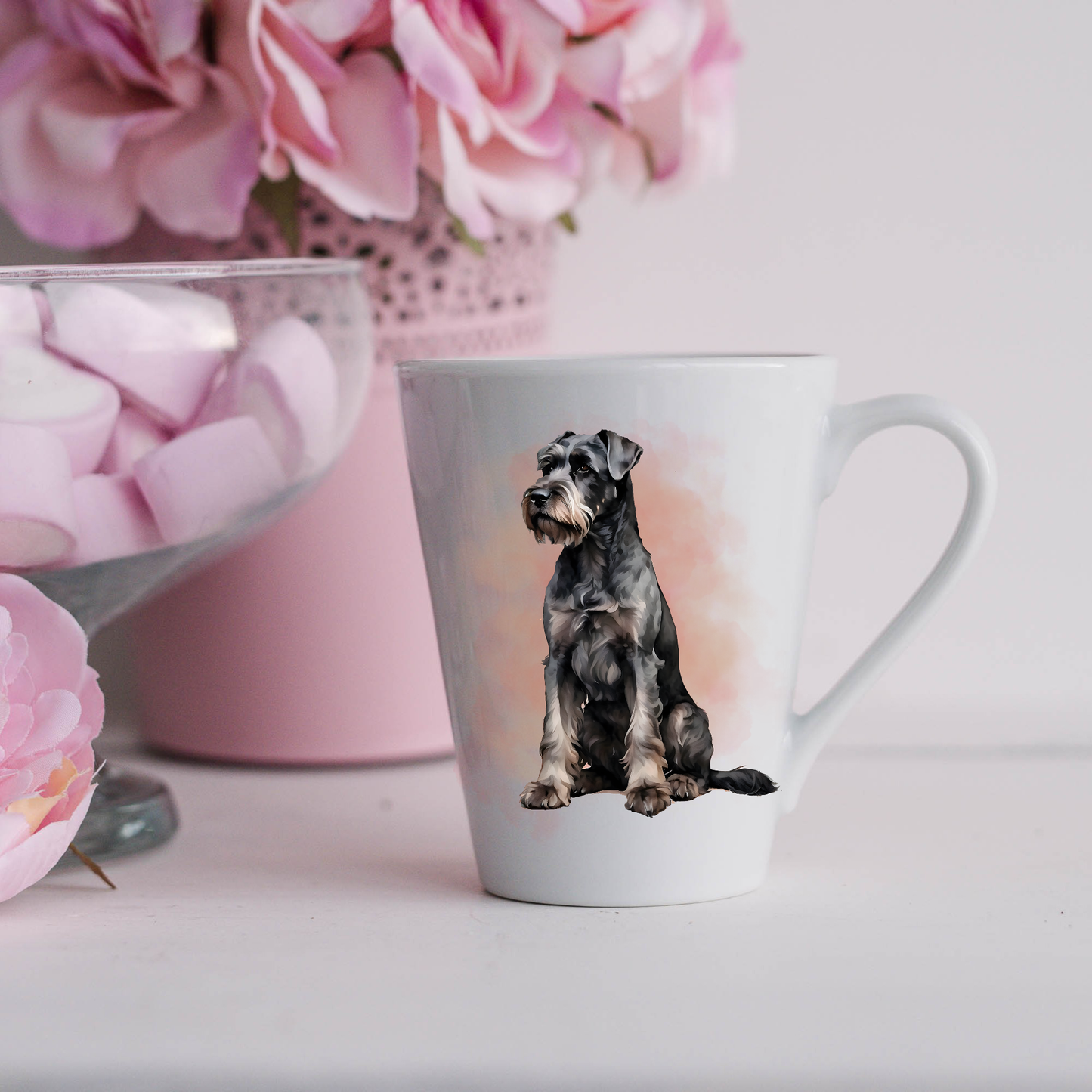 A Giant Schnauzer is shown sitting on a white ceramic latte mug in a smoky background and looking forward.  The mug is placed on a white surface with pink flowers in a pink plant pot and a dish of pink marshmallows.