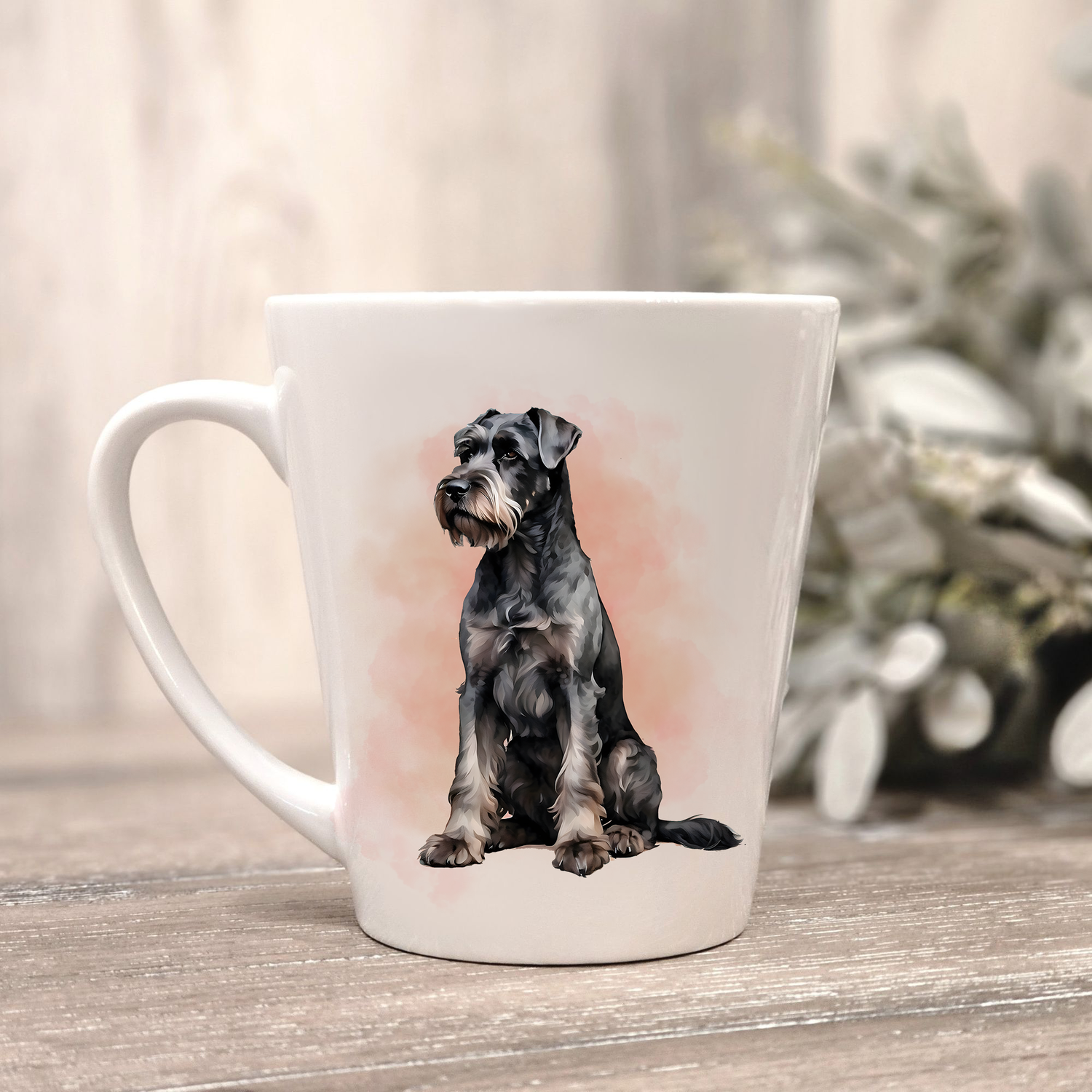 A Giant Schnauzer is shown in a sitting position on a white ceramic latte mug in a smoky background and looking forward.  The mug is placed on a wooden surface with sage green foliage to the right-hand side.