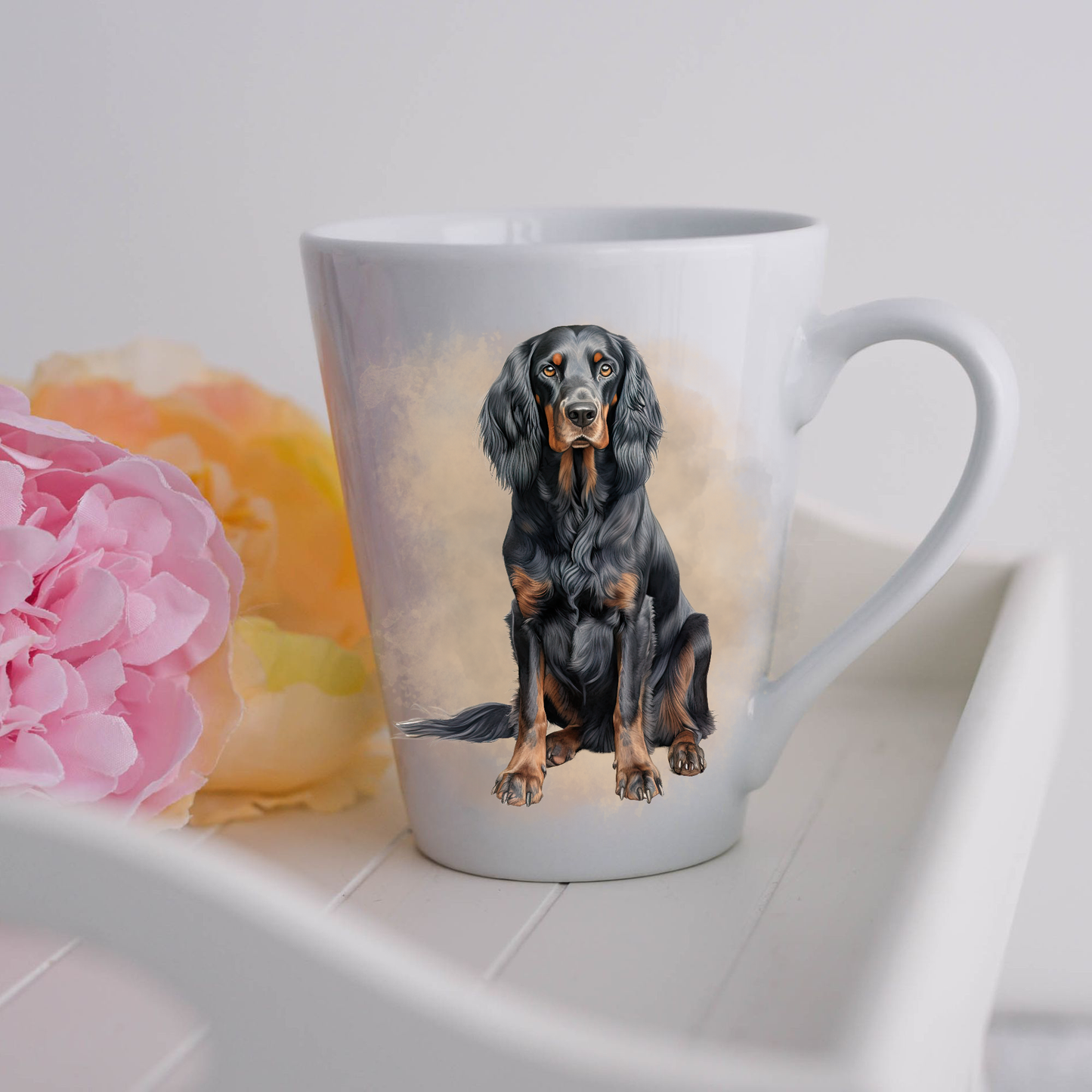 A Gordon Setter is in a seated position on a white ceramic latte mug in a smoky background and looking forward.  The mug is placed on a white tray with pink and yellow flowers. 