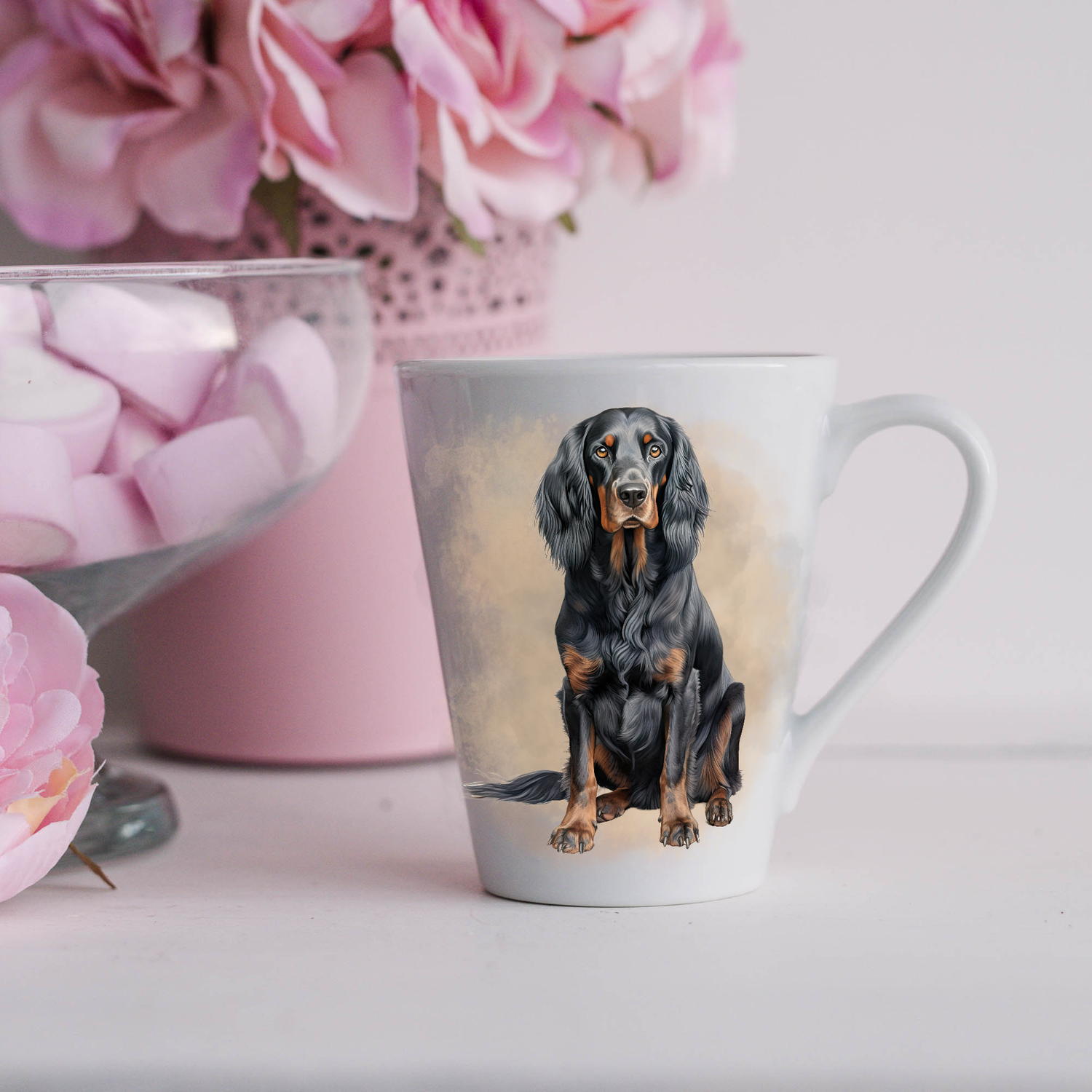 A black and tan Gordon Setter is displayed on a white ceramic latte mug in a smoky background and looking forward.  The mug is placed on a white surface with pink flowers in a pink plant pot and a dish of pink marshmallows.