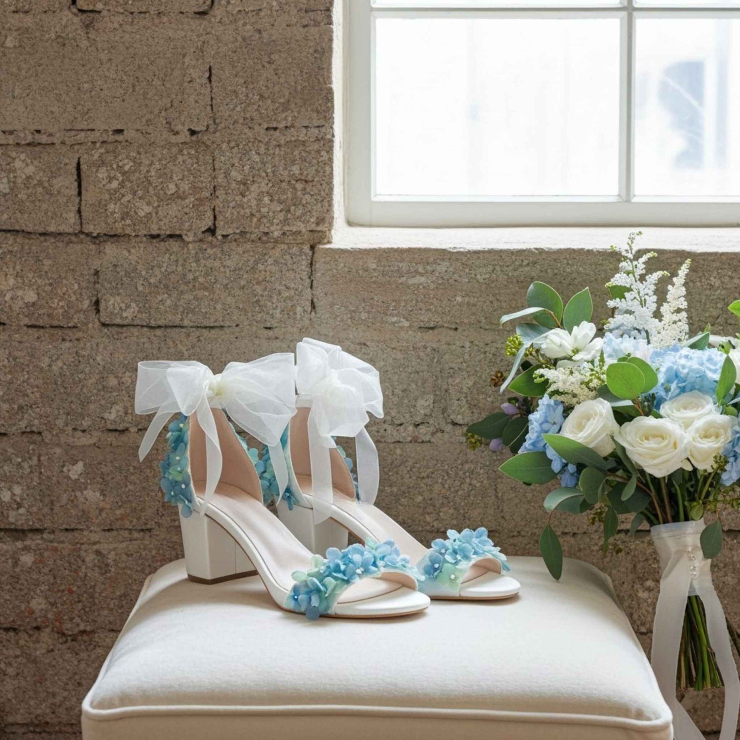 Ivory wedding shoes with blue floral decorations on a white cushion against a stone wall.