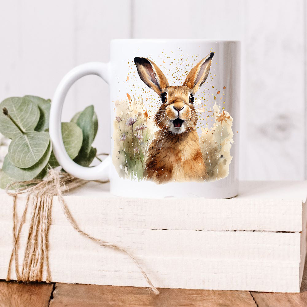 A smiley brown Spring Hare sits facing forward among purple and orange spring wildflowers on a white ceramic mug.    The mug is placed on a white wooden box and green leaves decorate the box with rustic string.