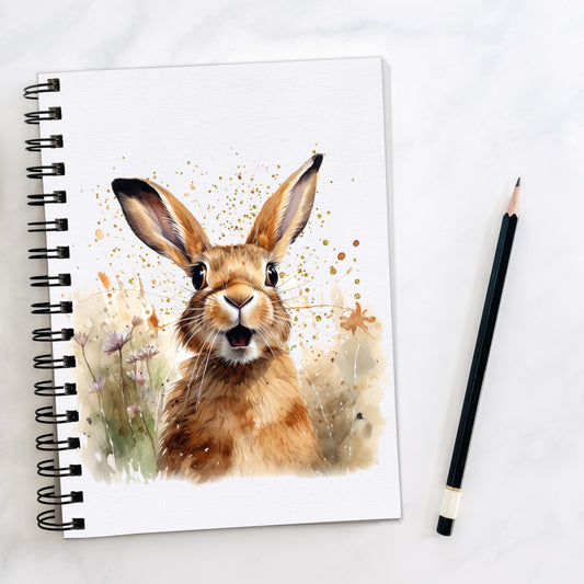 White spiral bound notebook, with a brown hare facing forward on a speckled background and sitting among flowers. A pencil is laid to the side.