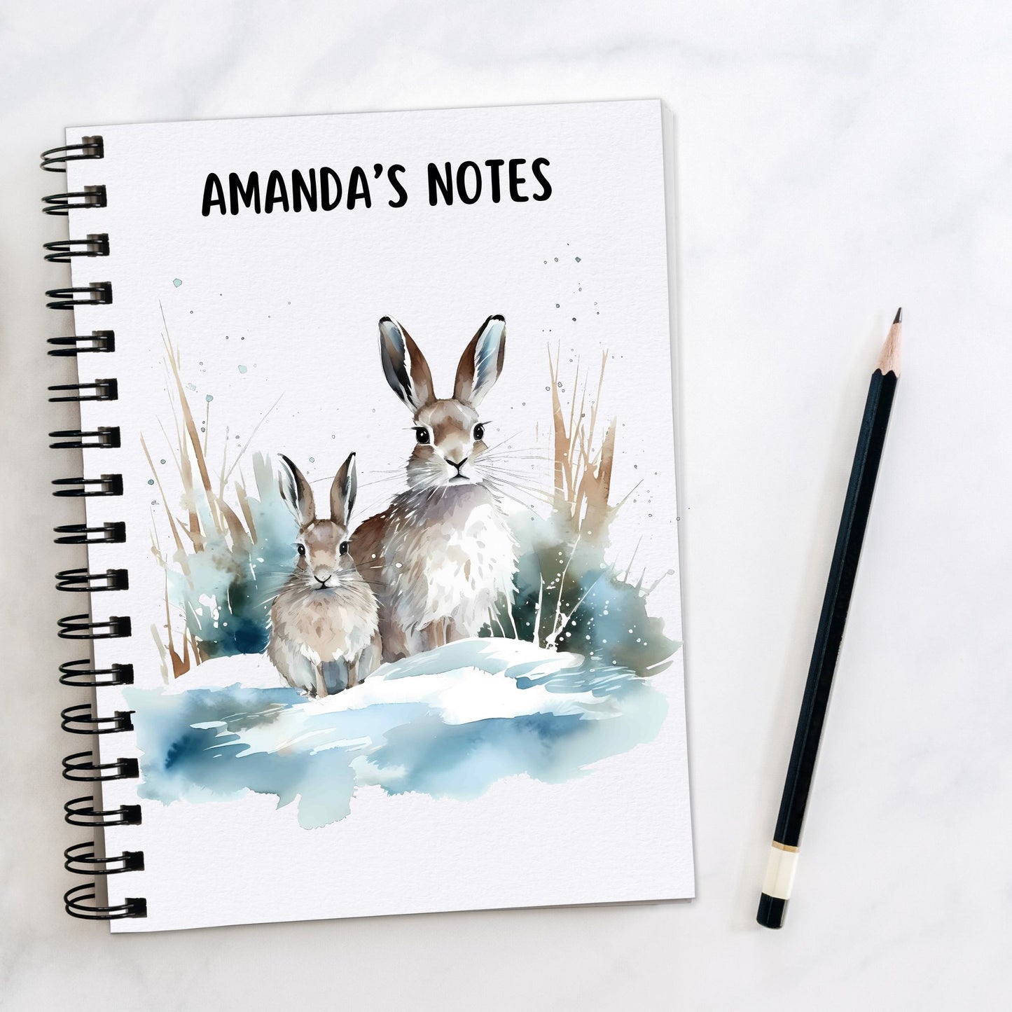 White notebook, personalised with a name. Two winter Hares are sat in a snowy scene. A pencil is laid to the side.