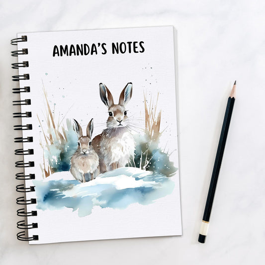 White notebook, personalised with a name. Two winter Hares are sat in a snowy scene. A pencil is laid to the side.
