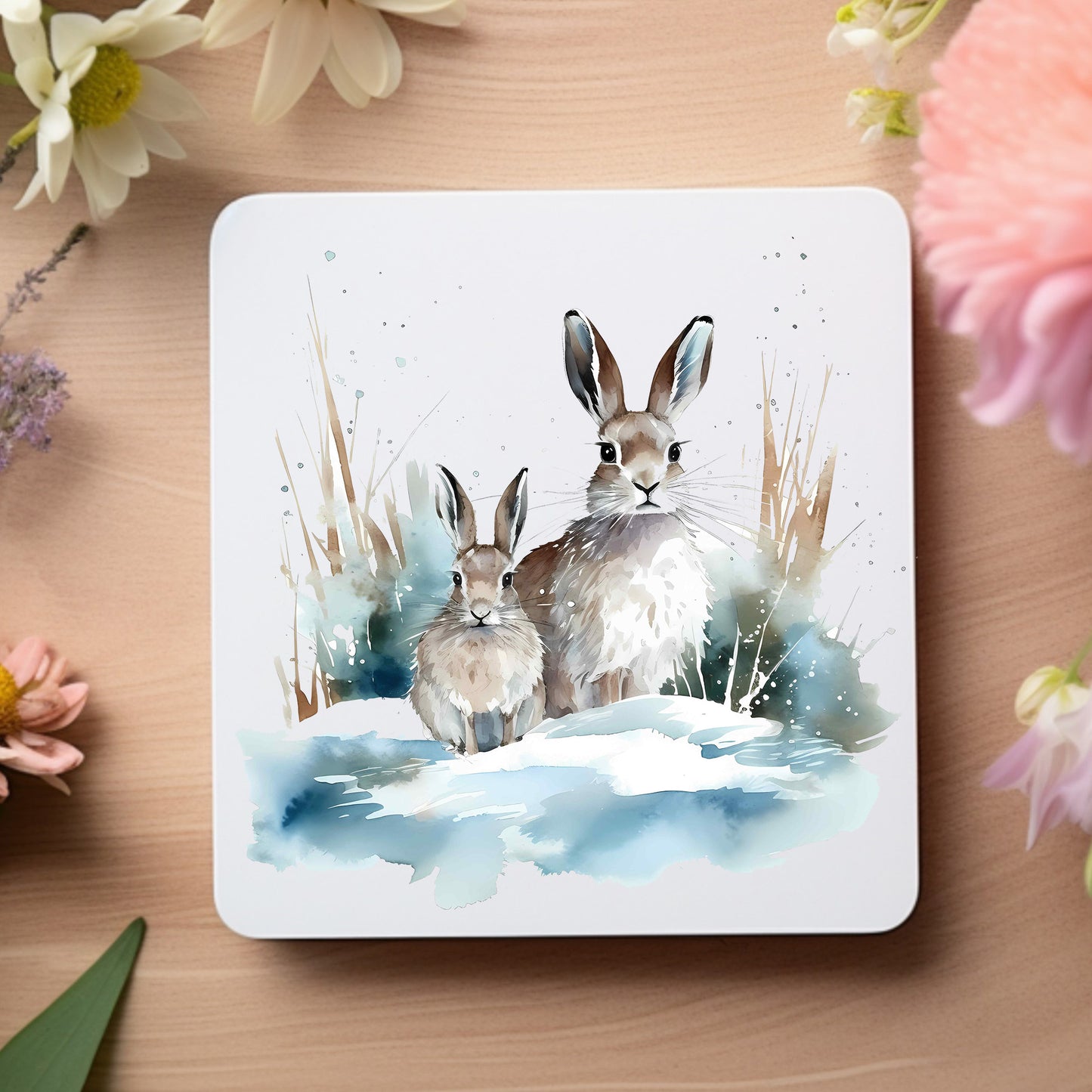 Two hares sat side by side in a snowy scene on a white coaster which is placed wooden surface with floral decoration.  