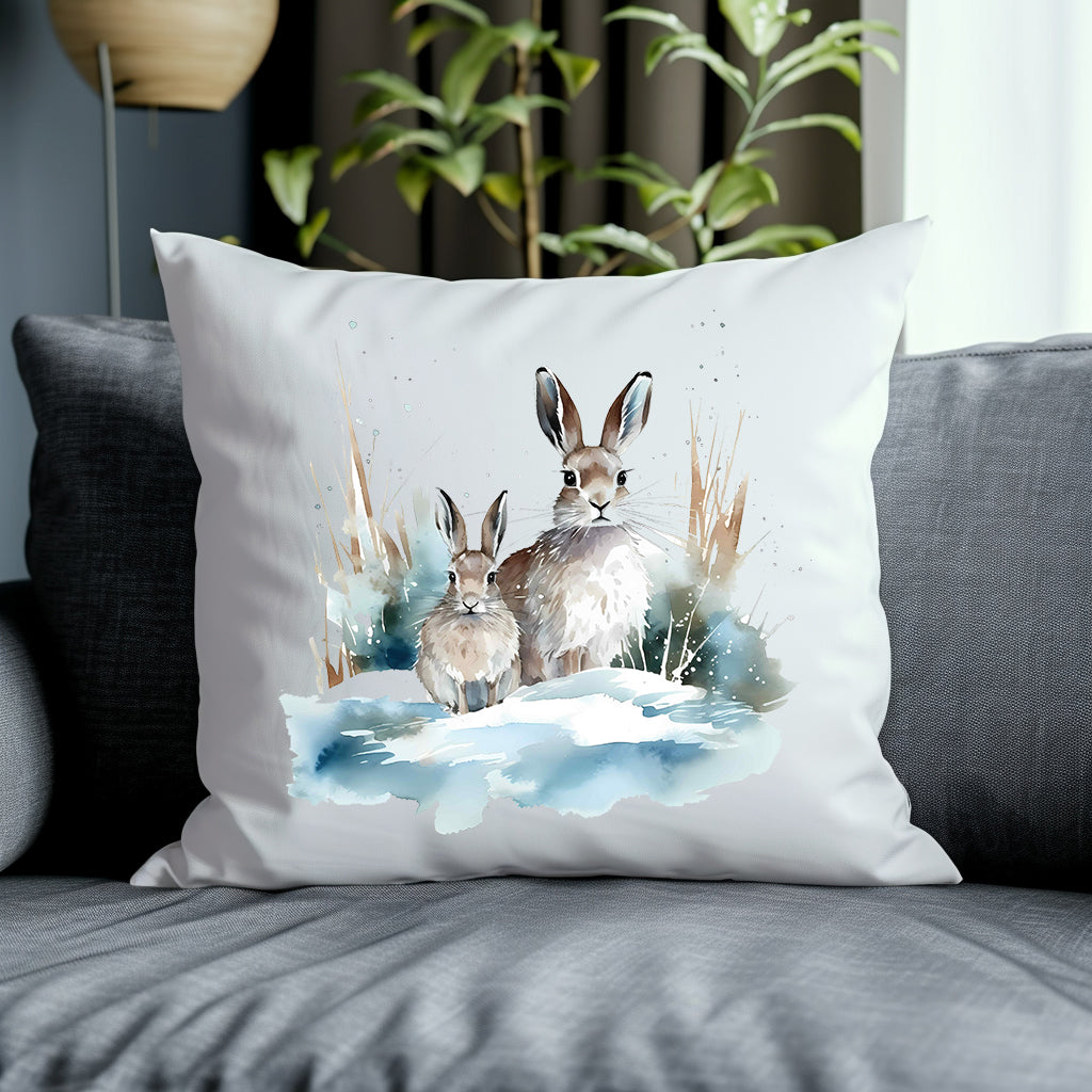 Two hares in a winter scene on a white cushion placed on a grey sofa with a green plant in the background