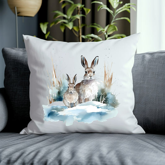 Two hares in a winter scene on a white cushion placed on a grey sofa with a green plant in the background