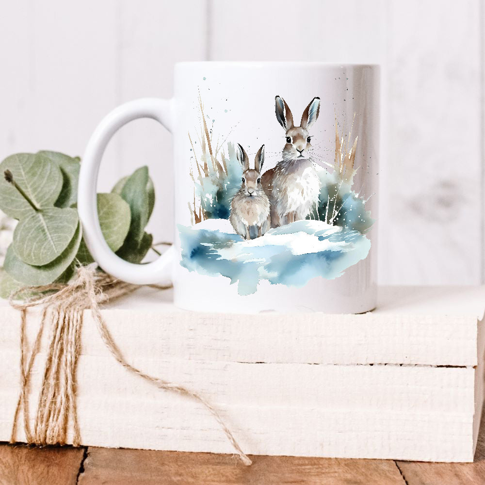 Two hares sat side by side in a snowy scene on a white ceramic mug which is placed on a white wooden box.  Green leaves decorate the box.