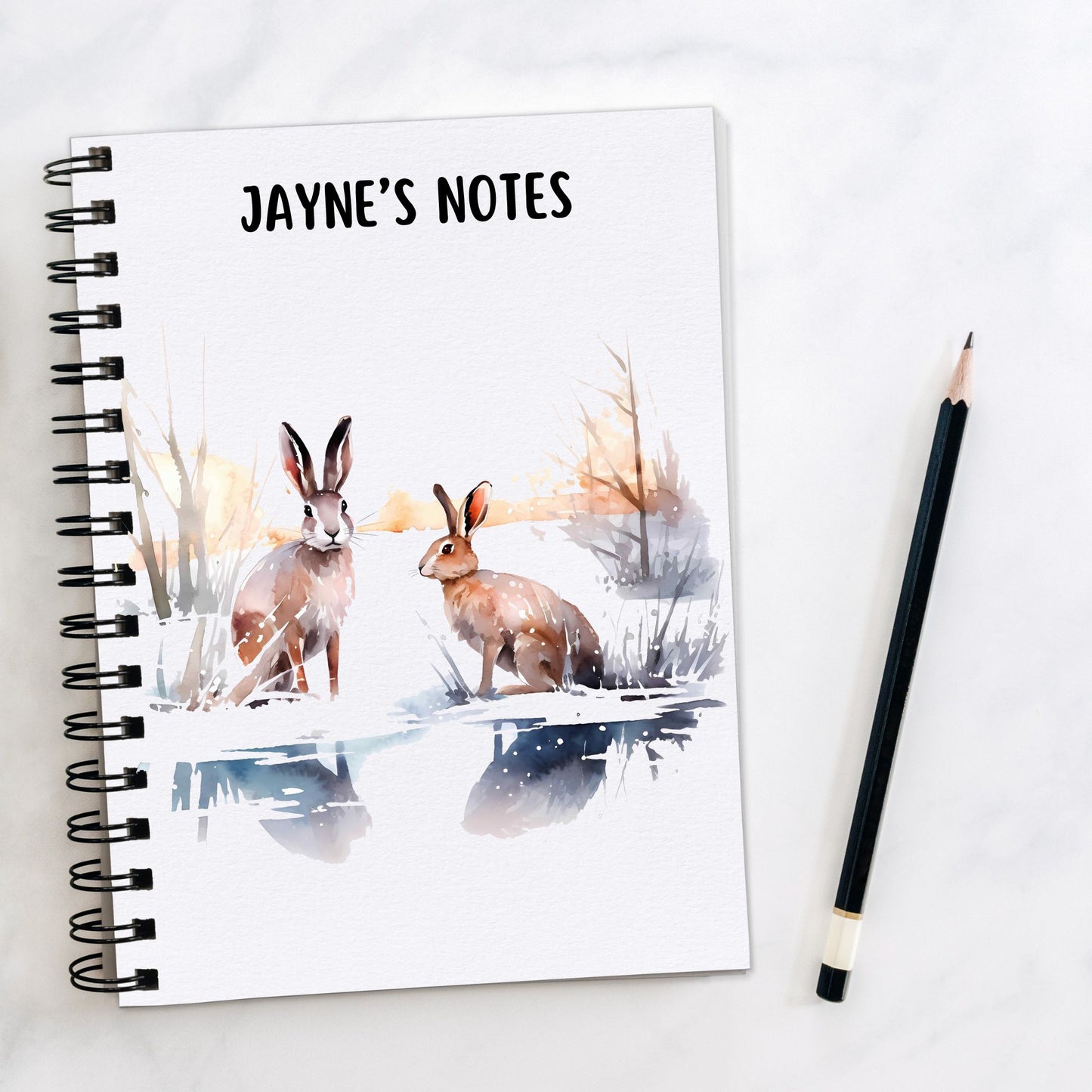 White notebook, personalised with a name. Two brown Hares are sat in a winter scene. A pencil is laid to the side.