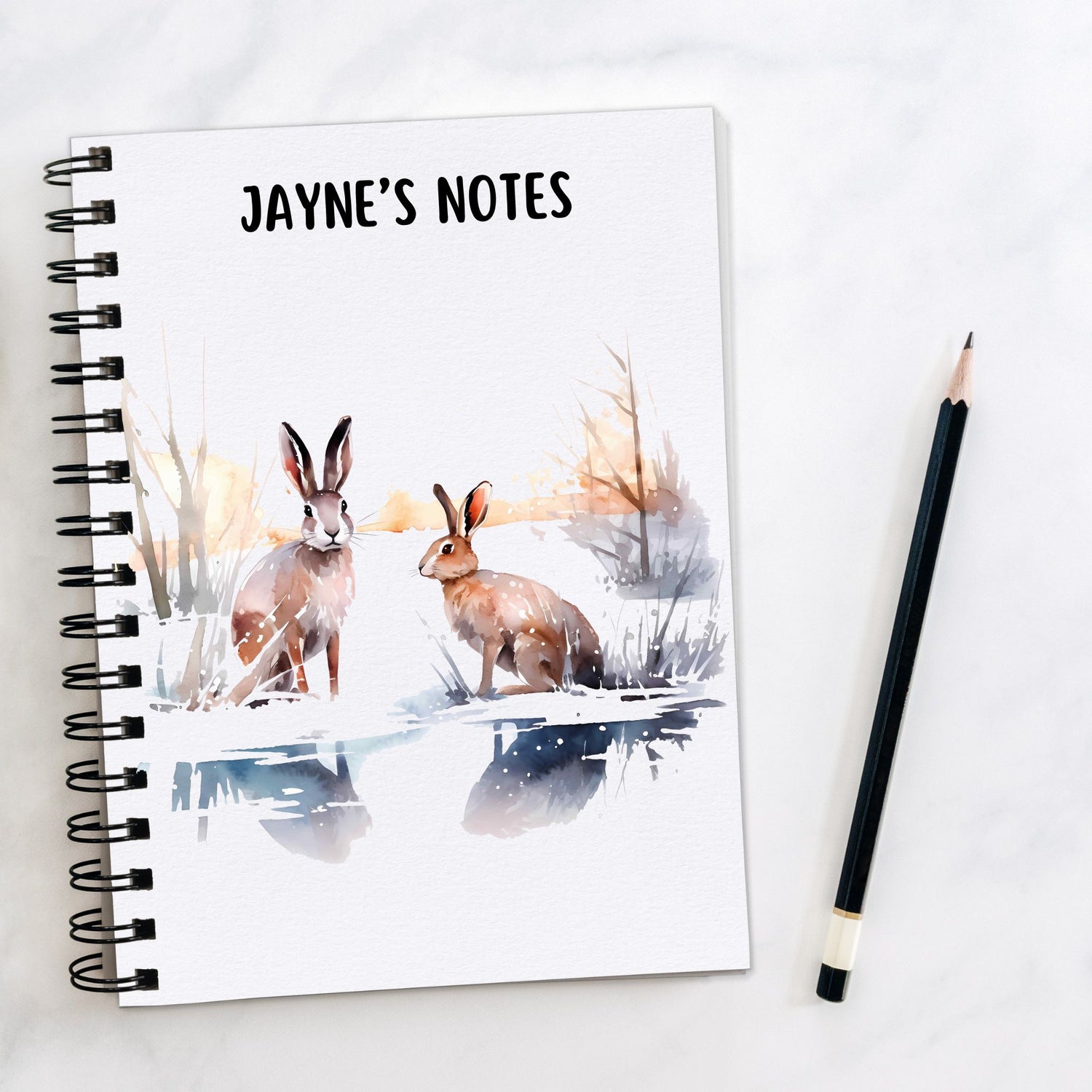 White notebook, personalised with a name. Two brown Hares are sat in a winter scene. A pencil is laid to the side.