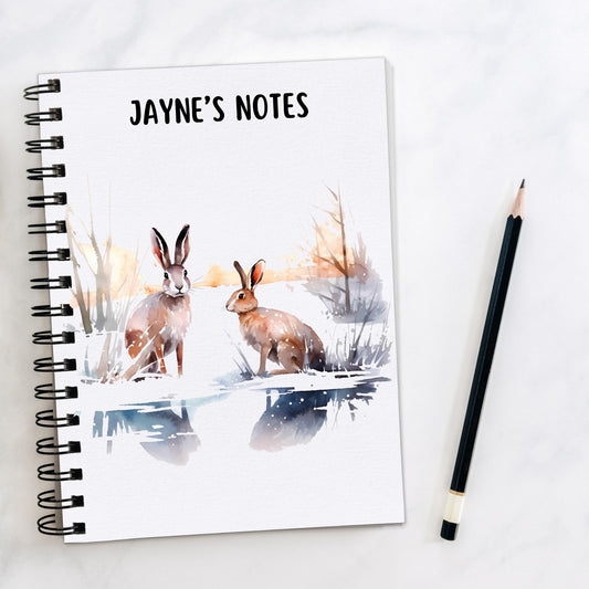 White notebook, personalised with a name. Two brown Hares are sat in a winter scene. A pencil is laid to the side.