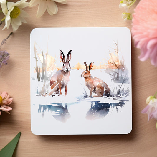 Two brown hares sat in a snowy scene on a white coaster which is placed on a wooden surface and floral decoration.