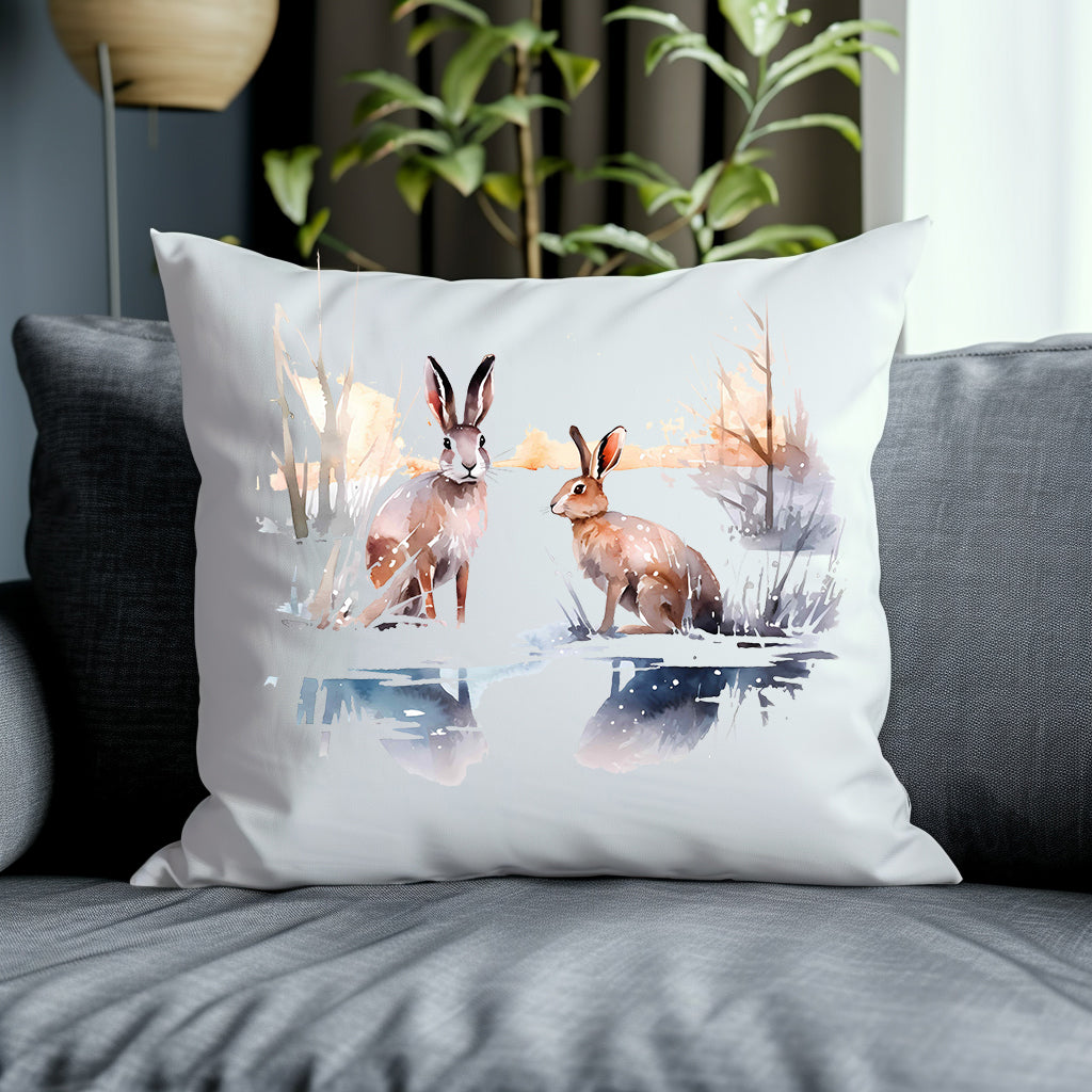 Two brown hares in a winter scene on a white cushion placed on a grey sofa with a green plant in the background