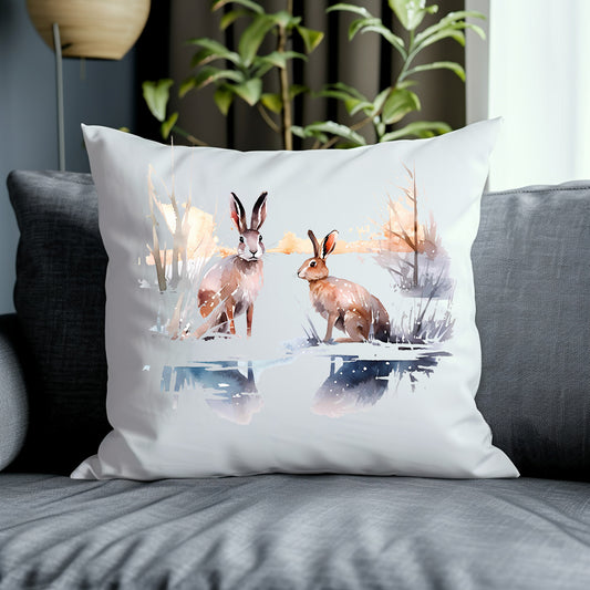 Two brown hares in a winter scene on a white cushion placed on a grey sofa with a green plant in the background