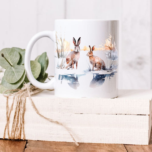 A pair of Winter Hares sit together in a snowy winter scene with their images reflected and bare trees in the background.  The mug is placed on a white wooden box and green leaves decorate the box with rustic string.