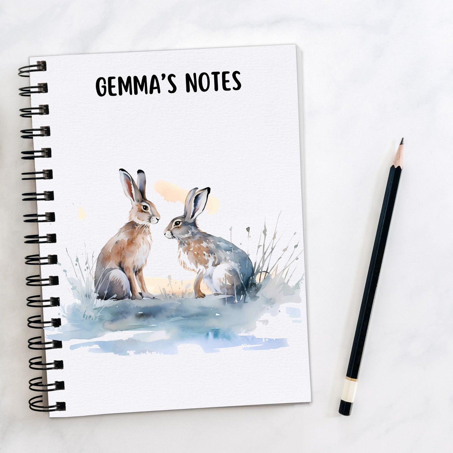 A personalised White notebook with two of Hares facing each other on a blue/grey surface with grass.  A pencil is laid to the side.