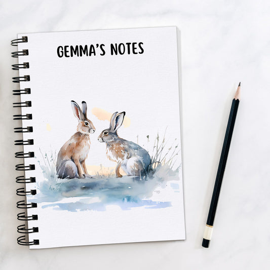 A personalised White notebook with two of Hares facing each other on a blue/grey surface with grass. A pencil is laid to the side.