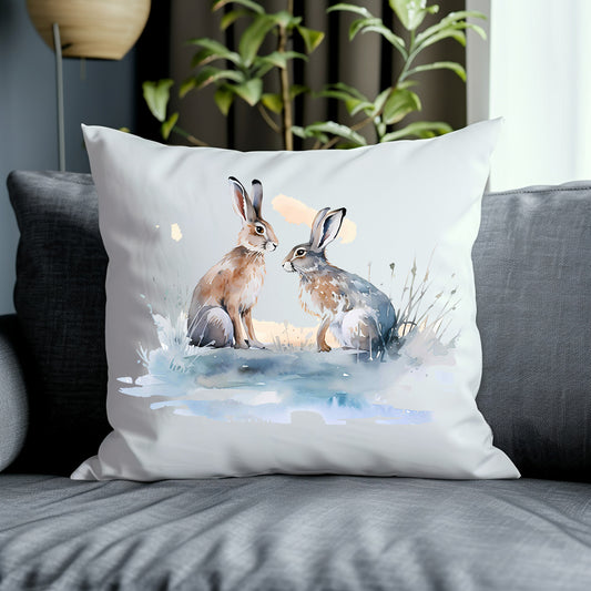 Two Hares face one another in a snow scene on the front of a white cushion which is placed on a grey sofa.  A green plant decorates the background.