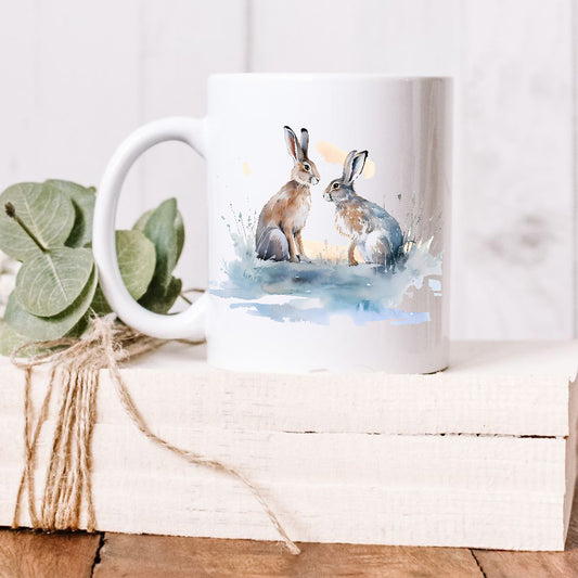 Two Winter Hares sit facing each other in a snow scene on a white ceramic mug.  The mug is placed on a white wooden box and green leaves decorate the box with rustic string