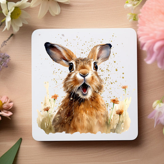 A brown Spring Hare with a surprised expression sits among orange spring wildflowers on a white coaster. The coaster is placed on a wooden surface flowers decorating the outer edge.