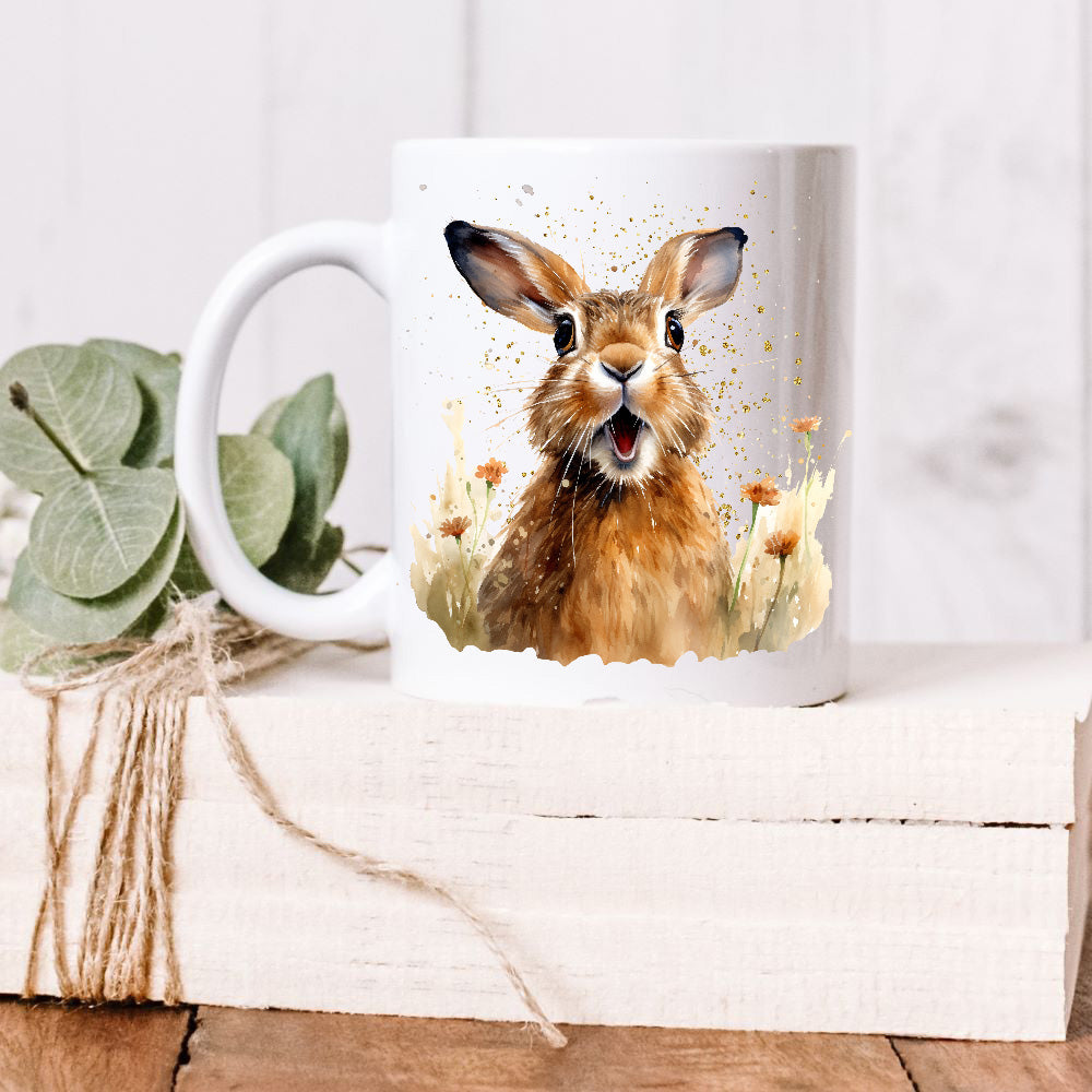 A brown Spring Hare with a suprised expression sits facing forward among orange wildflowers on a white ceramic mug.    The mug is placed on a white wooden box and green leaves decorate the box with rustic string.