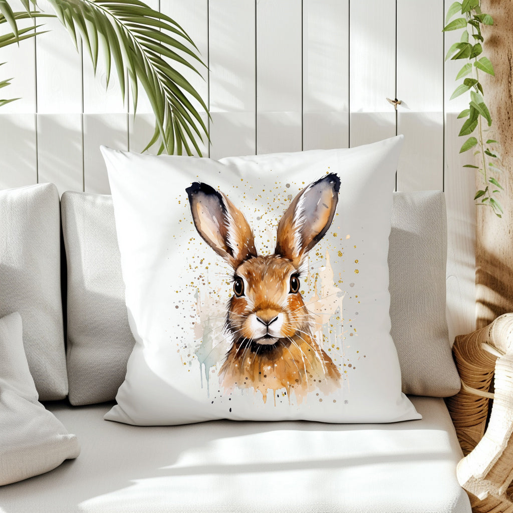A Spring Hare head and shoulders image with a splattered background on the front of a white cushion which is placed on a cream sofa.  White panels and green fern create the background.