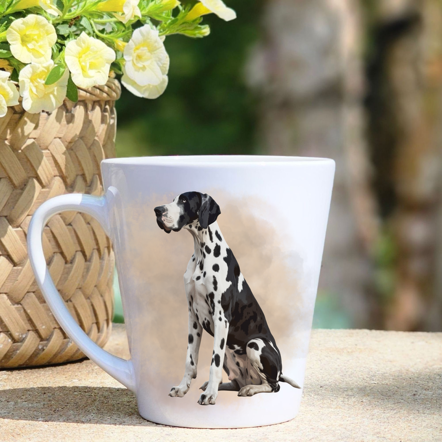 A white ceramic latte mug featuring a seated Harlequin Great Dane in a beige mist background.  A basket containing yellow flowers sits to the left-hand side.