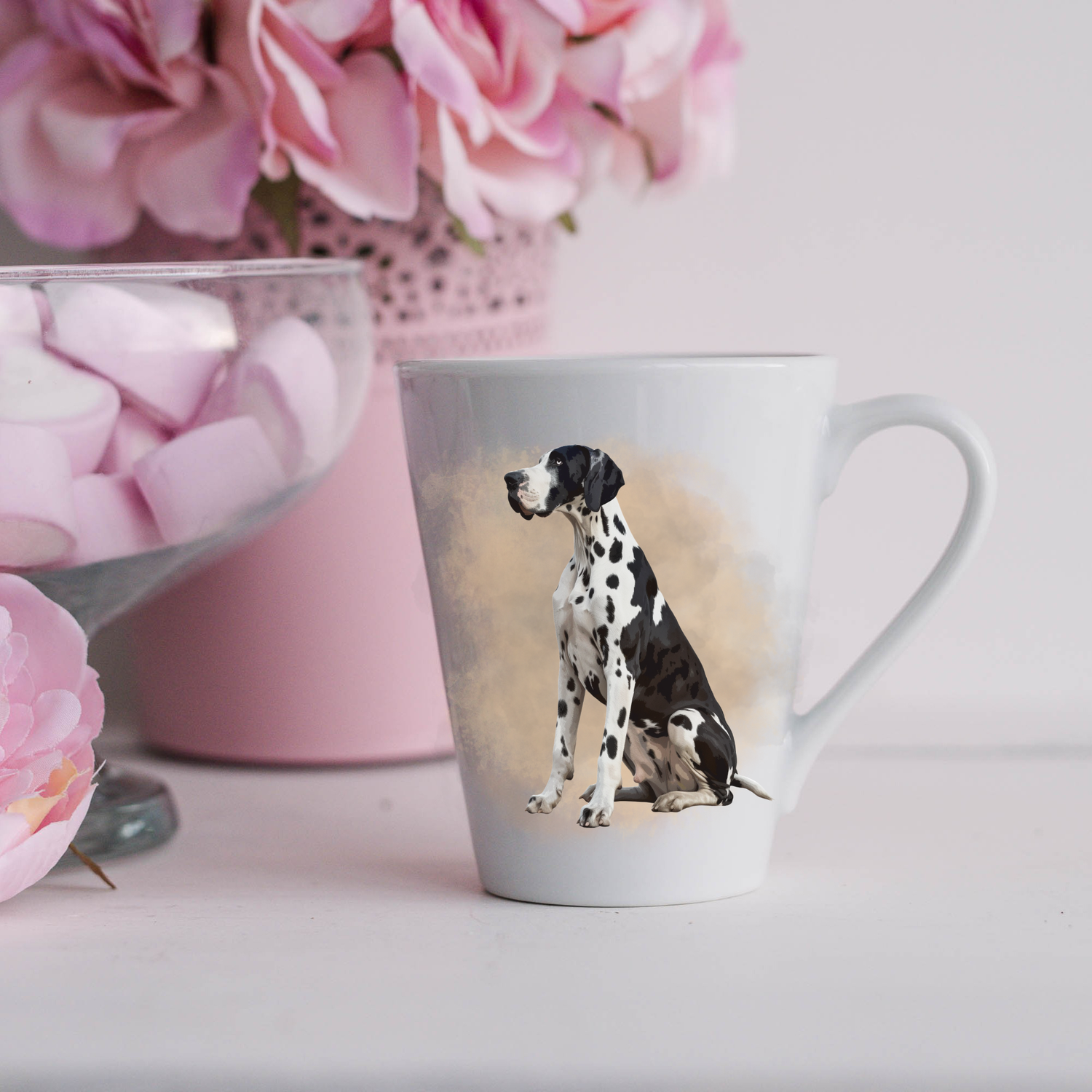A Harlequin Great Dane is seated in a smoky background on a white ceramic latte mug.  The mug is placed on a white surface with pink flowers in a pink plant pot and a dish of pink marshmallows.