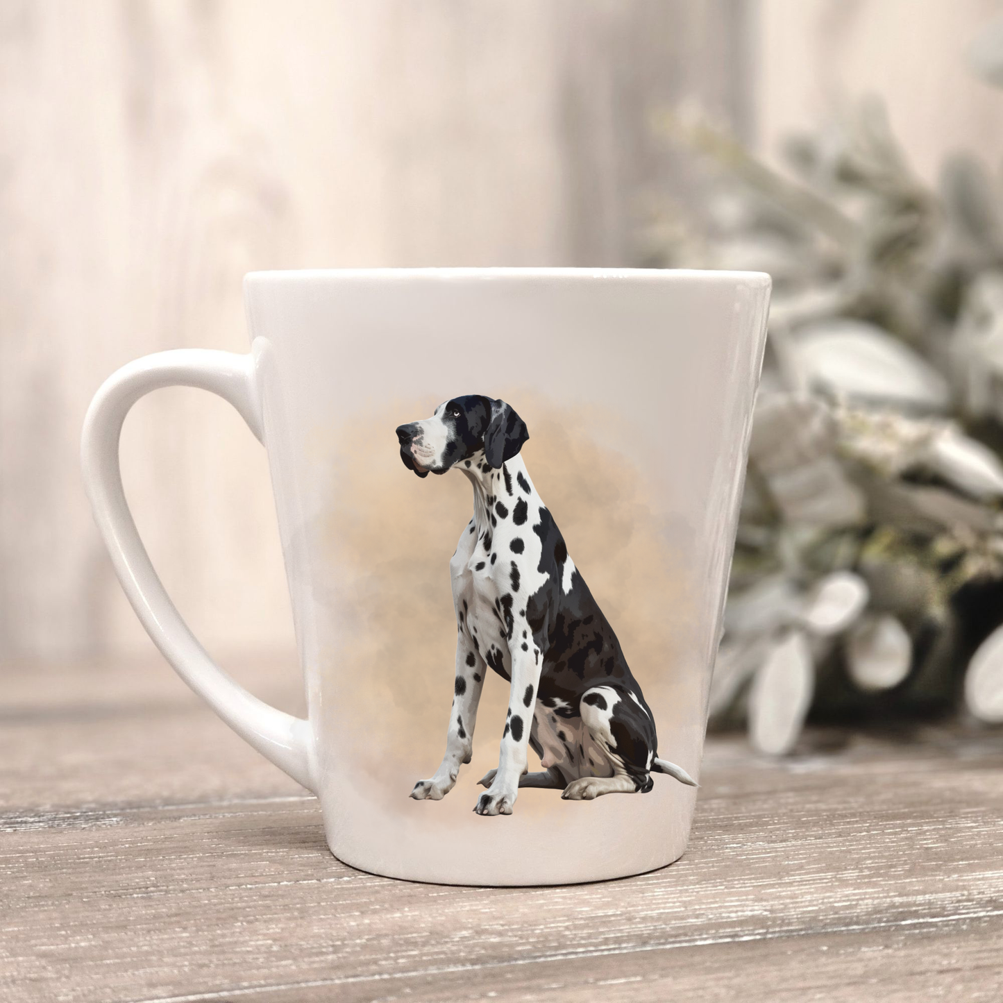 A Harlequin Great Dane is sitting facing left on a white ceramic latte mug in a smoky background and looking forward.  The mug is placed on a wooden surface with sage green foliage to the right-hand side.