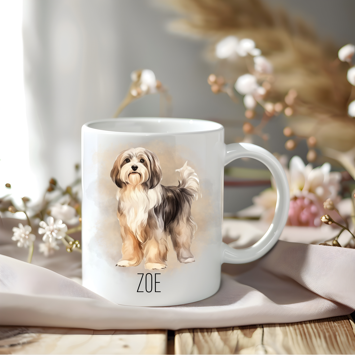 A Havanese coffee mug personalised with a pet name.  The mug is sat on a wooden surface with a creamy coloured cloth and various flowers adorn the background.