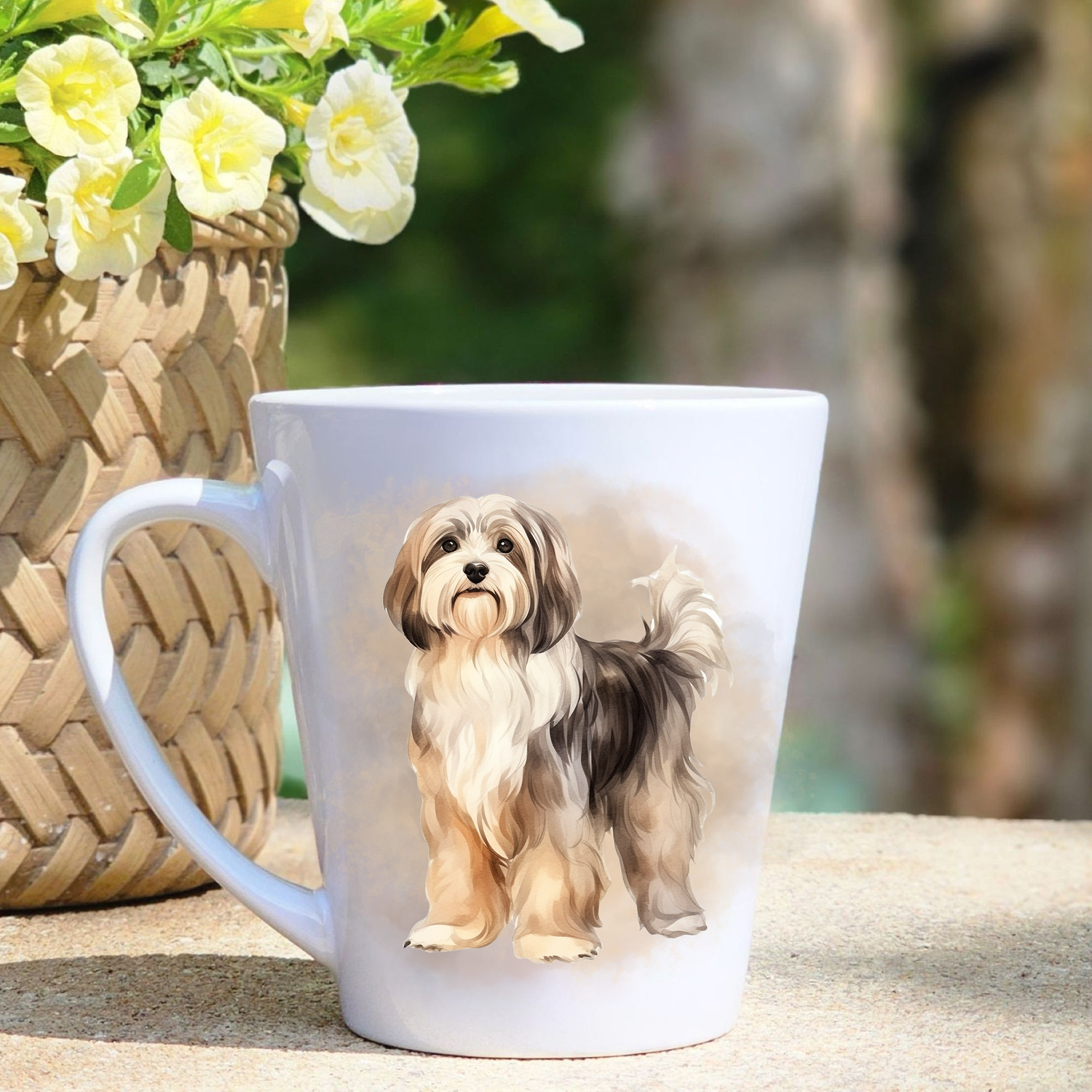 A white ceramic latte mug featuring a standing Havanese on a misty background.  A basket containing yellow flowers sits to the left-hand side.