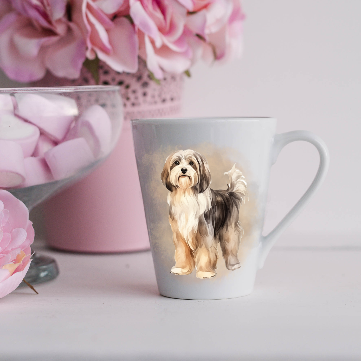 A cute Havanese dog is shown on a white ceramic latte mug in a smoky background and looking forward.  The mug is placed on a white surface with pink flowers in a pink plant pot and a dish of pink marshmallows.