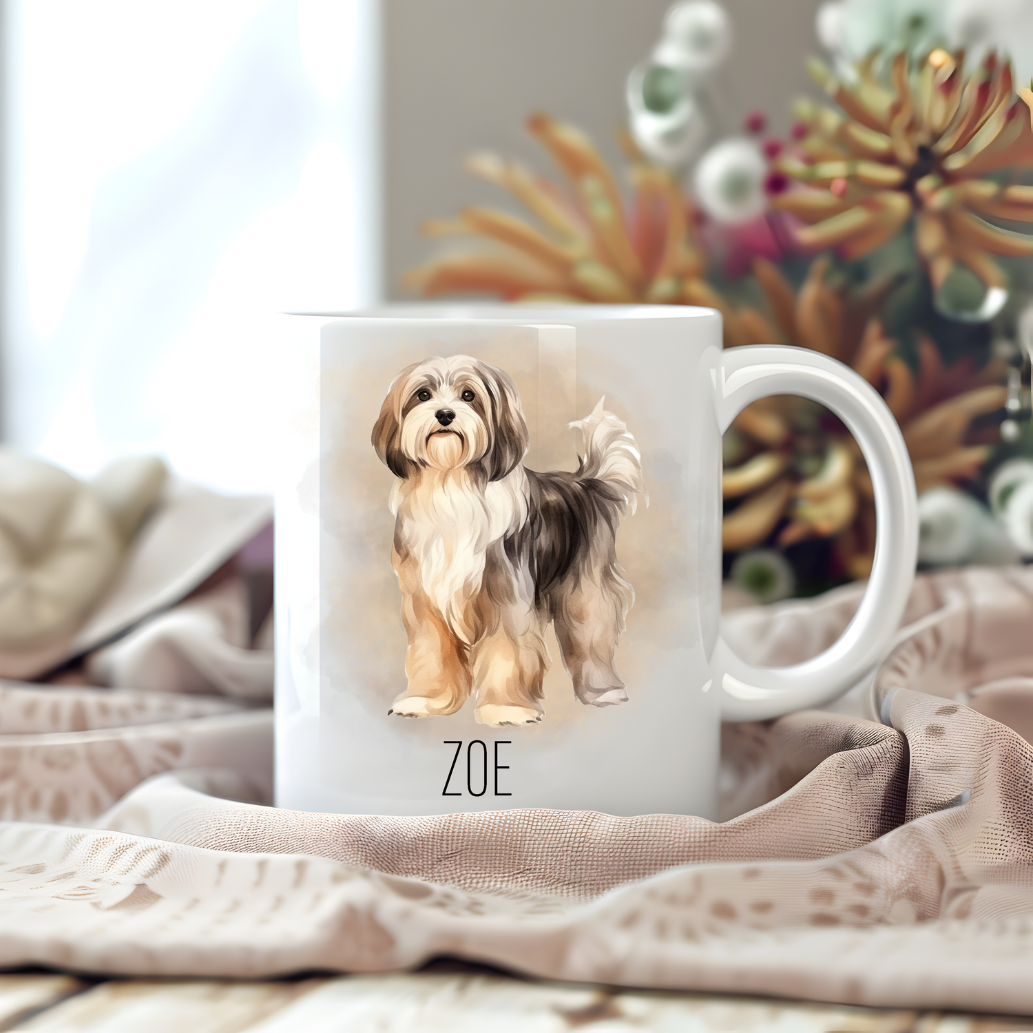 A personalised coffee mug featuring a Havanese dog on a white ceramic mug which has been placed on a beige cloth.  Stems of large  flowers accessorizes the image.