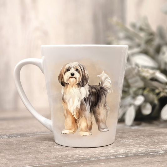 A Havanese dog is shown standing on a white ceramic latte mug in a smoky background and looking forward.  The mug is placed on a wooden surface with sage green foliage to the right-hand side.