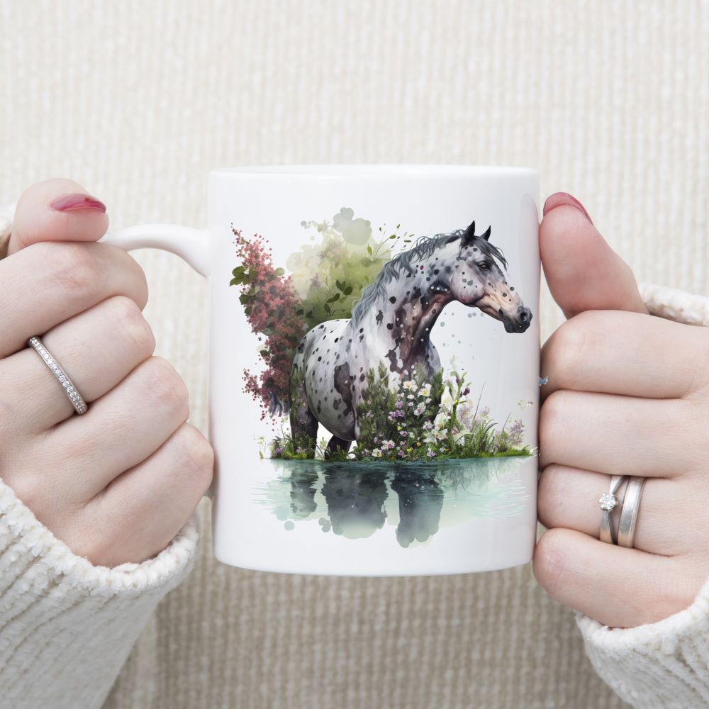 An Apaloosa horse standing in water with purple and white flowers decorating the image which is set on a white ceramic mug being held with both hands by a woman.