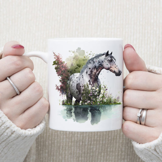 An Apaloosa horse standing in water with purple and white flowers decorating the image which is set on a white ceramic mug being held with both hands by a woman.