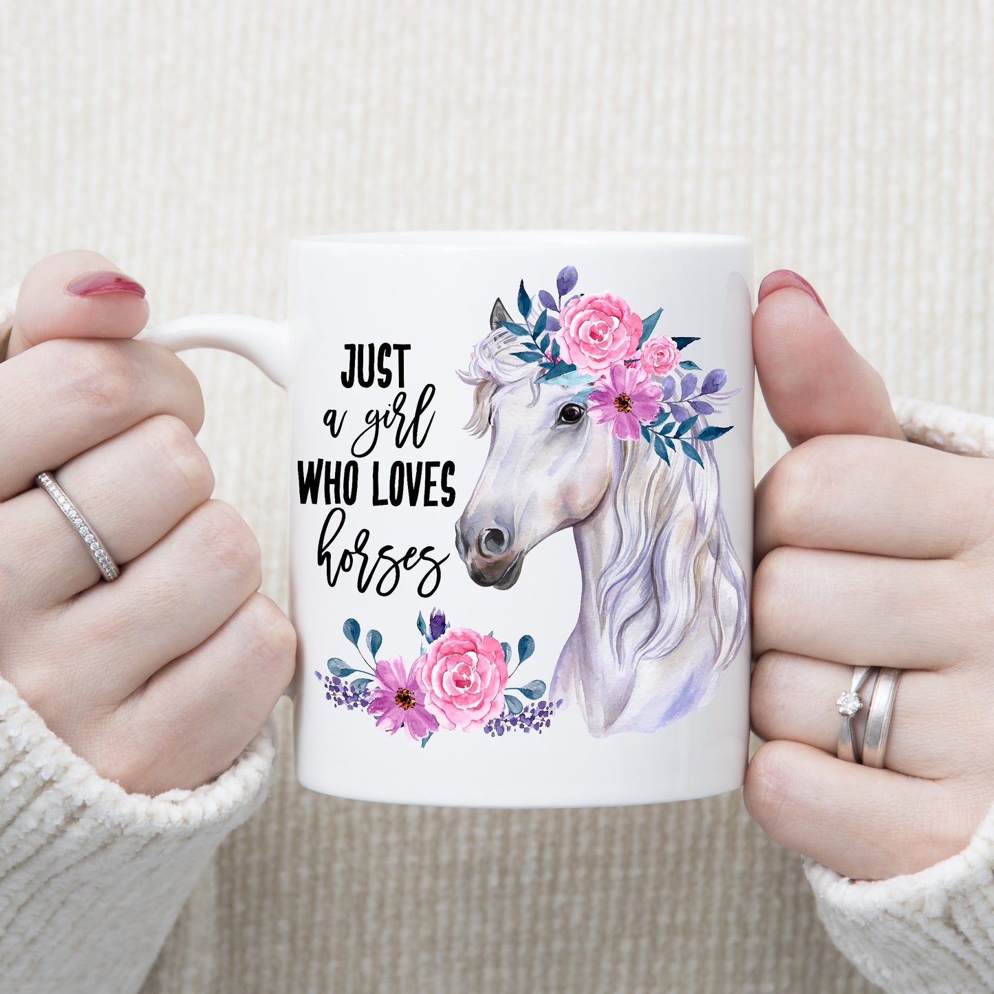 A head and shoulder watercolour image of a white horse with pink flowers in its mane. Image includes a quote 'Just a girl who loves horses' and is set on a white ceramic mug being held with both hands by a woman.