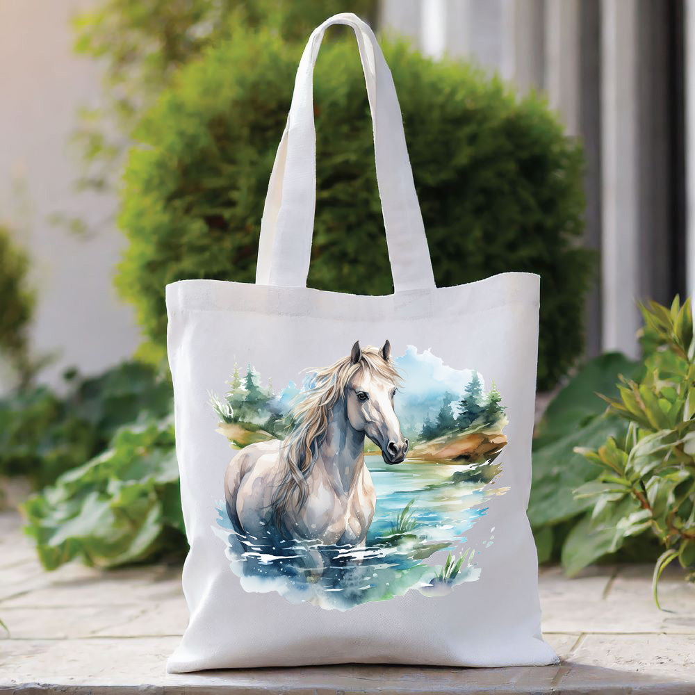White tote bag with a long handle showing a grey horse in a river scene with trees in the background.  The bag is placed in front of a large green shrub with green foliage to the right side.